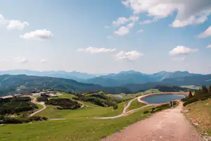 Campinganlage mit See, Uferweg und Wohngebäuden im Tal, umgeben von grünen Wiesen und Bergen unter blauem Himmel.