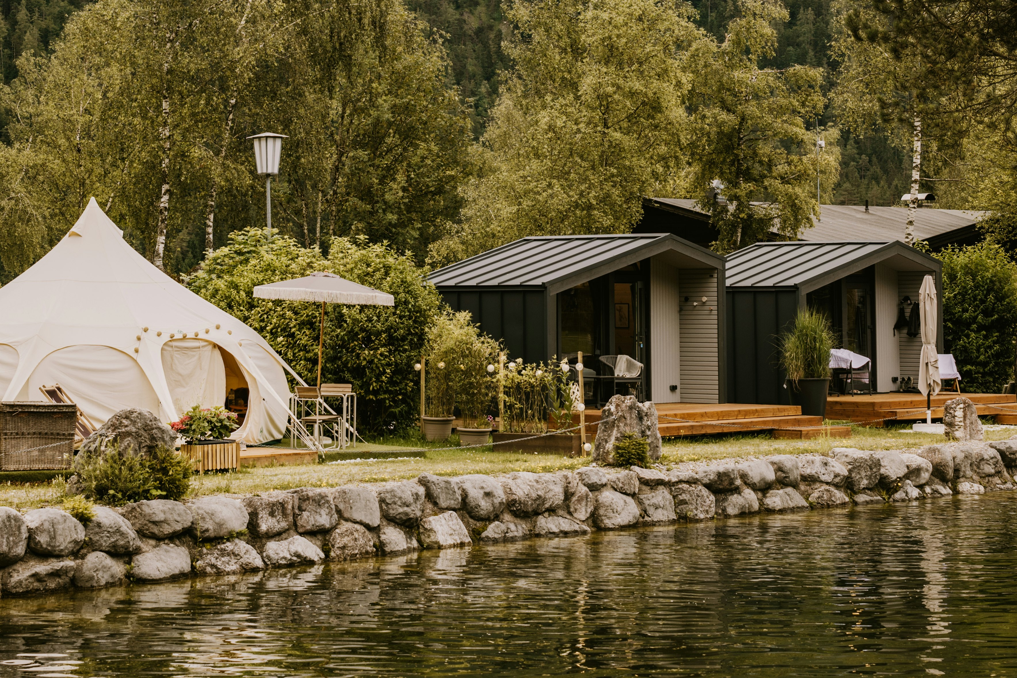 Weißes Zelt neben Holzbungalows am See, mit Terrasse, Stühlen, Tisch und Laternen im grünen Garten.