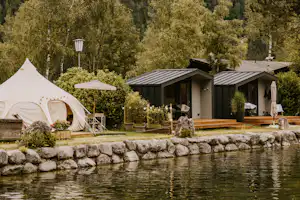 Weißes Zelt neben Holzbungalows am See, mit Terrasse, Stühlen, Tisch und Laternen im grünen Garten.