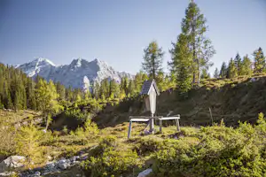 Bergpanorama mit kleinem Camping-Unterstand, Bank und Tisch auf einer Wiese, umgeben von Lärchen und Felsgestein.