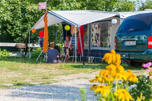Campingzelt mit Lichterkette und Sitzbereich, daneben Wohnwagen und Auto, im Vordergrund gelbe Blumen im Garten.