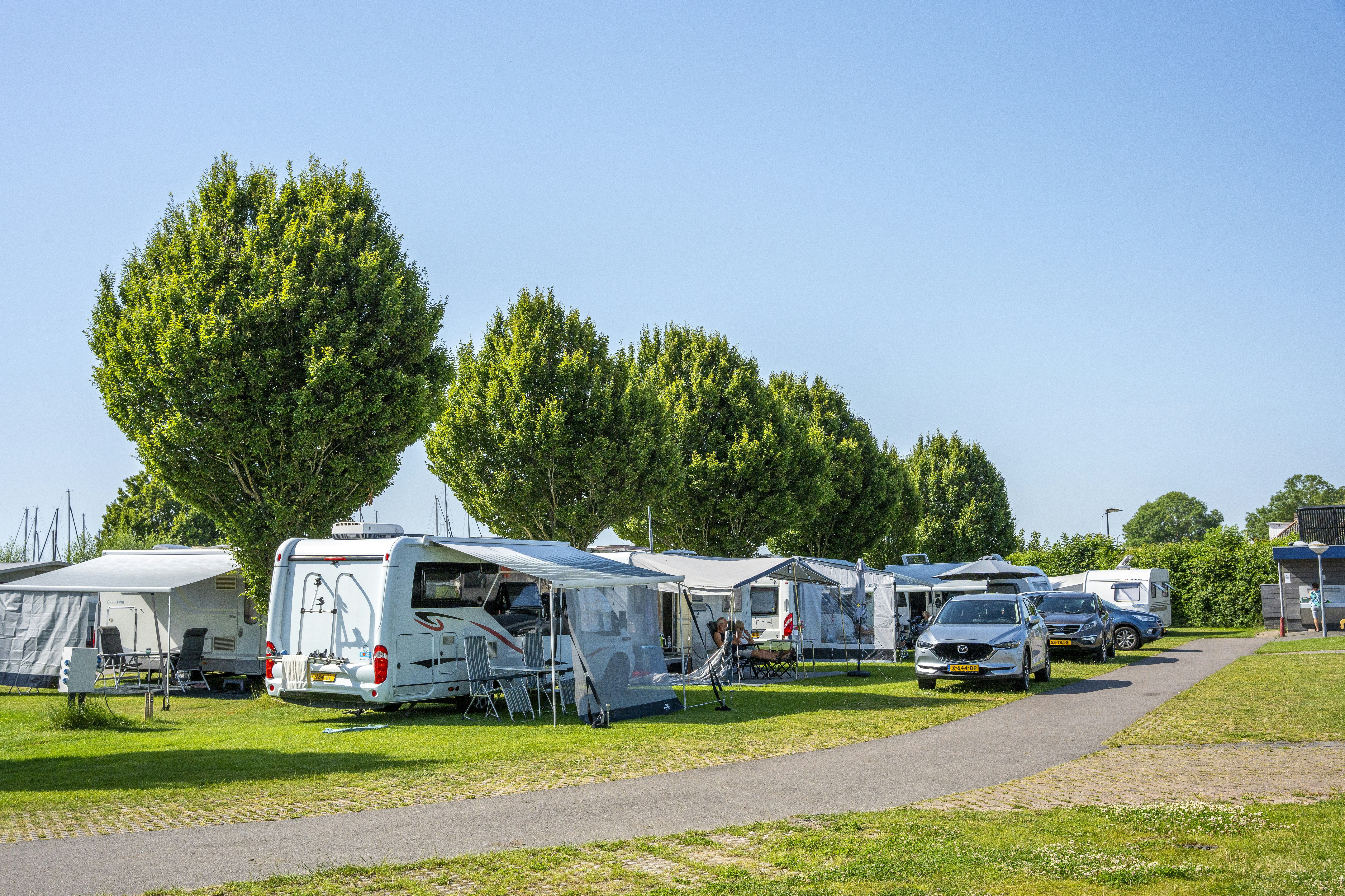 Reihe von Wohnmobilen und Wohnwagen auf einem Campingplatz, mit Vorzelten, Rasenflächen und einem breiten Weg zwischen den Stellplätzen.