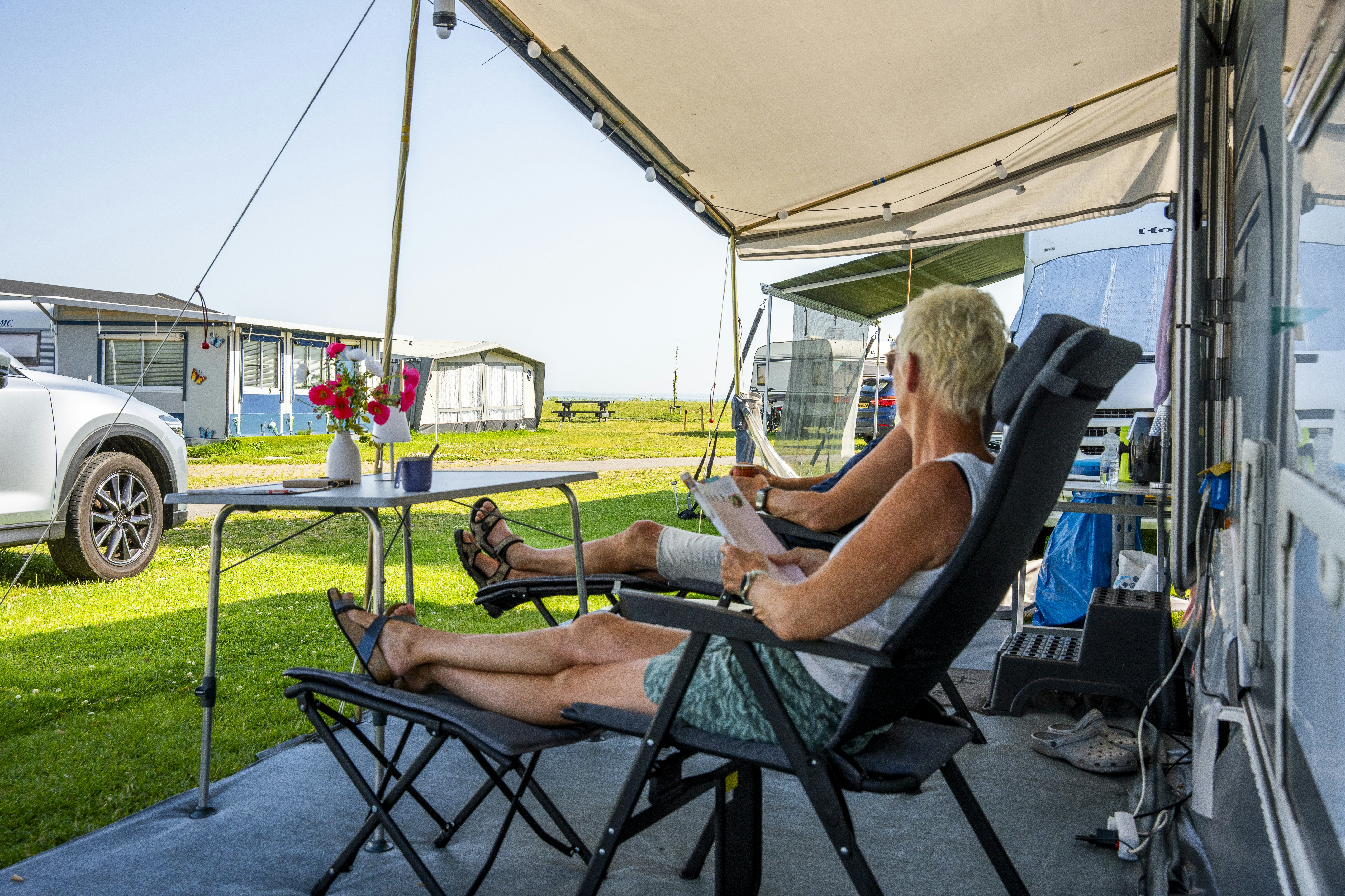 Campinggast sitzt auf einem Klappstuhl unter dem Zeltvorzelt und liest am Wohnwagen, mit Rasen und Blumen im Hintergrund.