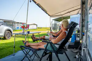 Campinggast sitzt auf einem Klappstuhl unter dem Zeltvorzelt und liest am Wohnwagen, mit Rasen und Blumen im Hintergrund.