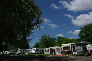 Campingplatz mit Wohnmobilen und Zelten, Sitzgelegenheiten und Bäumen vor blauem Himmel mit Wolken.