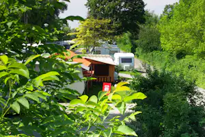 Wohnwagen mit Markise und Terrasse im Grünen, umgeben von Bäumen und Hecken auf einem Campingplatz.