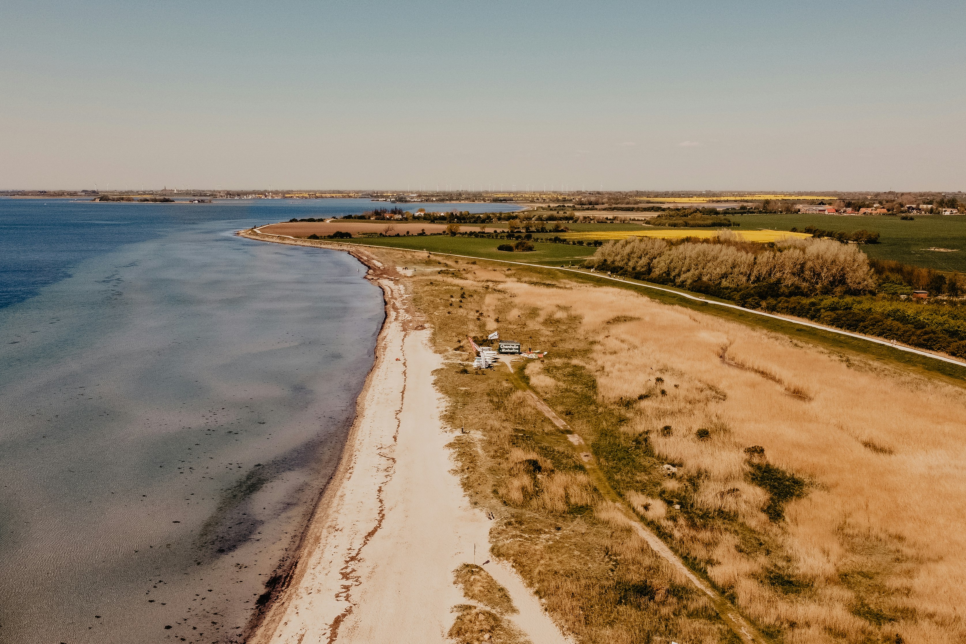 Küstenlinie mit Strand und Uferweg, Blick auf Felder und Häuser entlang der Bucht.