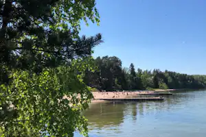 Seeufer mit Steg und vielen Badegästen, im Vordergrund grüne Bäume und ruhiges Wasser unter klarem Himmel.