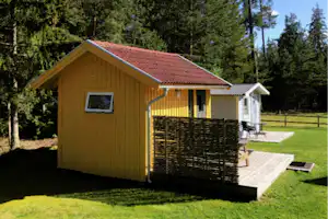 Gelbes Holzhaus mit rotem Ziegeldach, Terrasse mit Sichtschutz und Sitzbank, umgeben von Wiesen und Bäumen im Campingbereich.