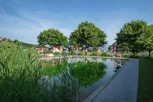 Teich am Campingplatz mit gepflastertem Weg, Bäumen und roten Sonnenschirmen am Ufer.