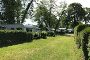 Wohnwagen und Wohnmobil auf einem Campingplatz, umgeben von Hecken, Rasen und Bäumen, mit Auto am Stellplatz.