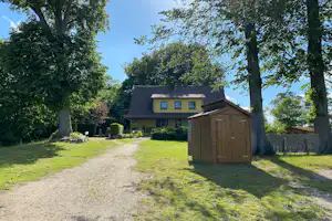 Weg am Campingplatz mit Rasen, Holzschuppen und einem Haus im Hintergrund zwischen großen Bäumen.