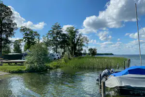 Zelt am Flussufer mit Wohnwagen und Bäumen im Hintergrund, grünes Gras und Steg am Wasser.