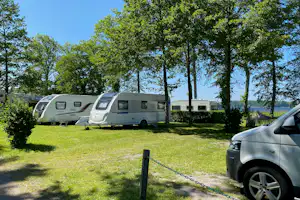 Wohnwagen auf einem Campingplatz-Stellplatz im Grünen, mit Bäumen, Grasfläche und geparktem Auto am Rand.