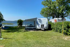Campingplatz am See mit Vorzelt und Wohnwagen auf einem Rasenplatz, Hecke und Bäume im Hintergrund.