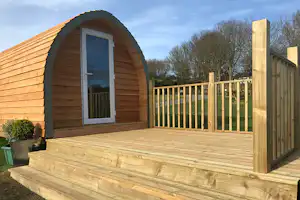 Holzbungalow mit halbrunder Tür und großer Holzterrasse, Geländer und Fensterfront, im Grünen am Rand eines Campingplatzes.