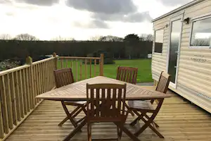 Wohnwagen mit Terrasse, Holztisch und Stühlen auf dem Campingplatz, Blick auf den Rasen und Bäume im Hintergrund.