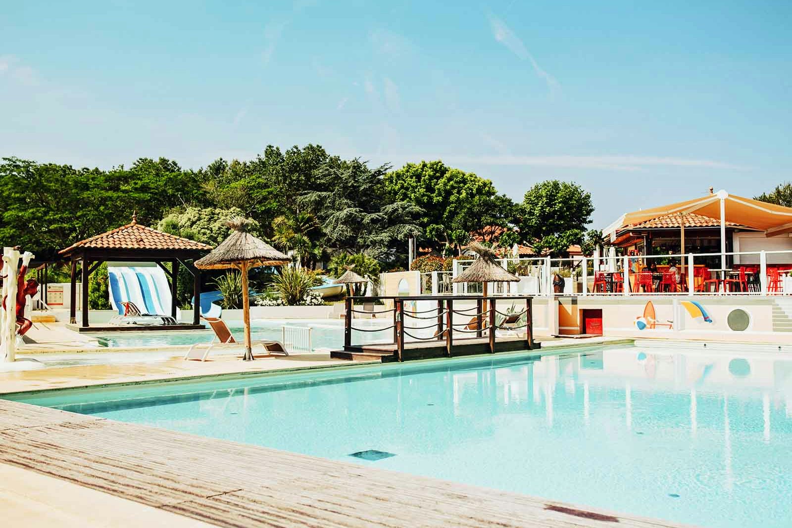 Schwimmbad auf dem Campingplatz mit Holzliegefläche, Sonnenschirm und Blick auf Bäume und Gebäude im Hintergrund.