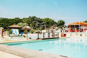 Schwimmbad auf dem Campingplatz mit Holzliegefläche, Sonnenschirm und Blick auf Bäume und Gebäude im Hintergrund.