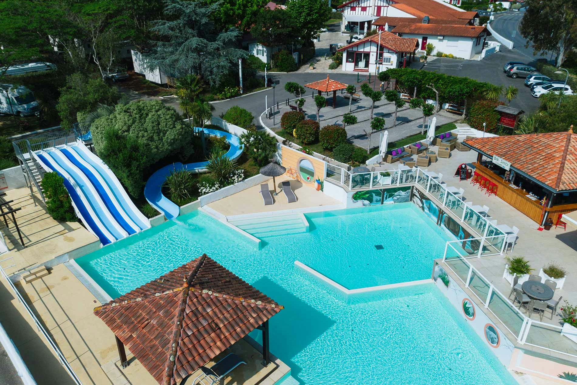 Großes Schwimmbecken mit Sonnendeck, daneben ein kleiner Pool und Liegen, im Hintergrund Gebäude und Bäume.
