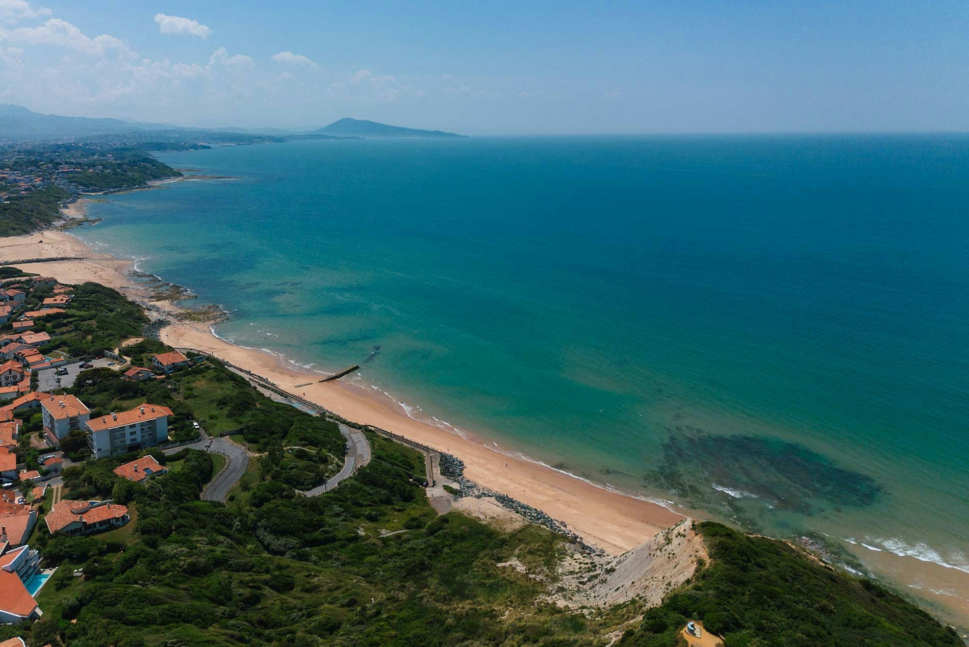 Luftaufnahme einer Küste mit Sandstrand, türkisfarbenem Meer, Häusern am Hang und einer Straße entlang der Bucht.