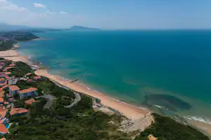 Luftaufnahme einer Küste mit Sandstrand, türkisfarbenem Meer, Häusern am Hang und einer Straße entlang der Bucht.