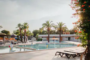 Camping-Pool mit Liegen am Beckenrand, Palmen im Hintergrund und ein weißes Gebäude hinter dem Poolbereich.