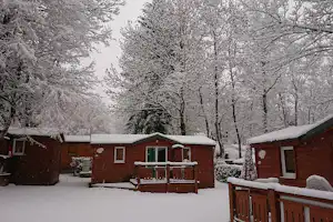 Holz-Campinghütten mit Veranda und Treppe im Schnee, umgeben von verschneiten Bäumen und Wegen.