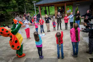 Kinder und Familien stehen auf dem Betonweg vor einem überdachten Bereich am Campingplatz, im Hintergrund ein grüner Hügel.