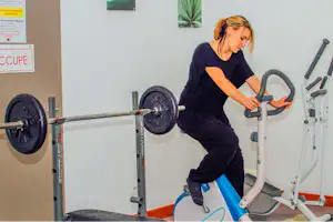 Frau nutzt ein Fitnessgerät im Innenbereich, neben Hantelscheiben und einer Langhantel im Trainingsraum.