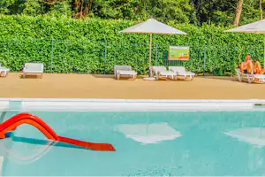 Camping-Pool mit roter Rutsche, Liegen und Sonnenschirmen vor einer hohen Hecke im Garten.