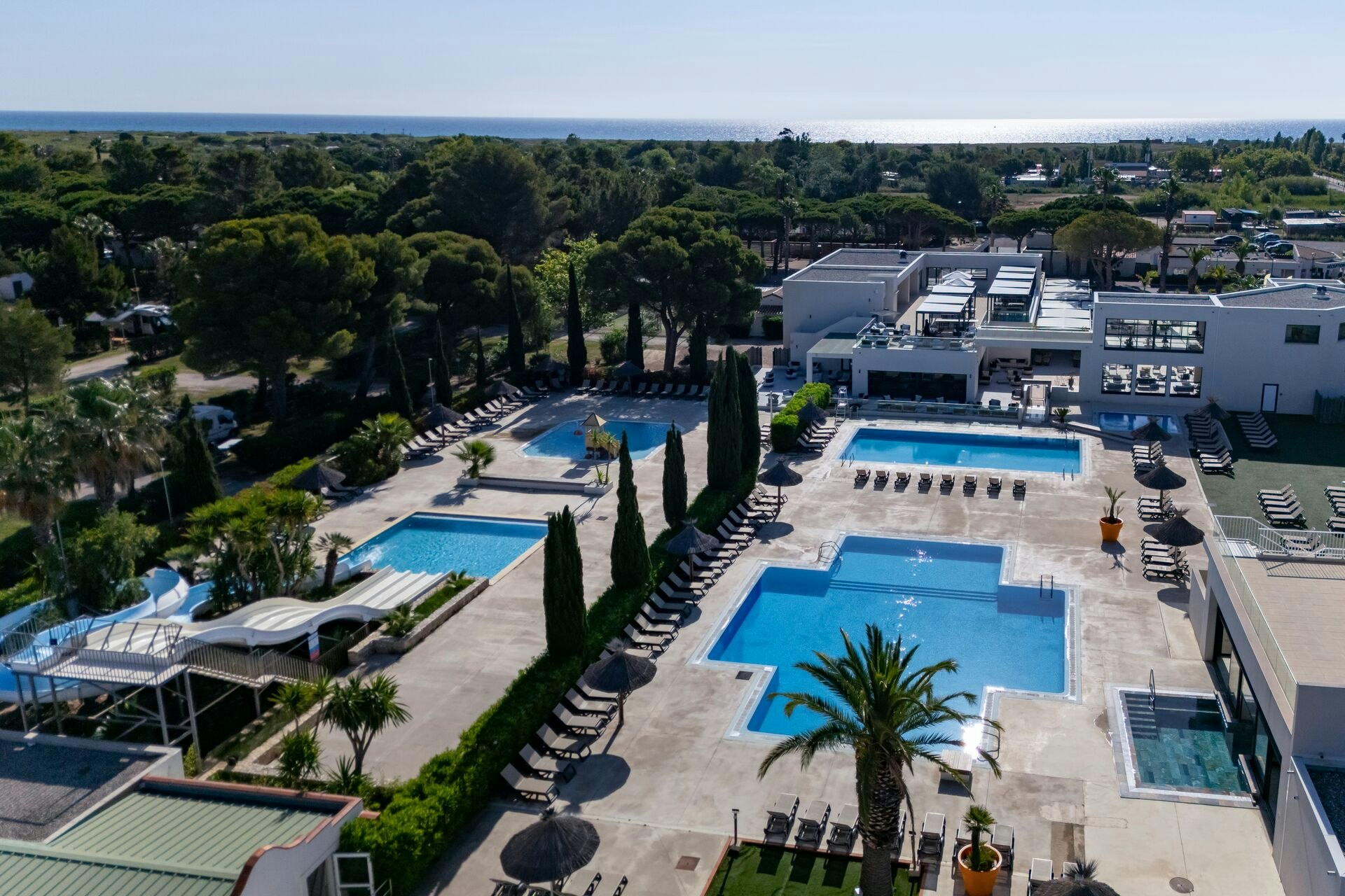 Blick auf mehrere Swimmingpools und Liegen am Campingplatz, umgeben von gepflegten Wegen, Palmen und Bäumen sowie weißen Gebäuden.