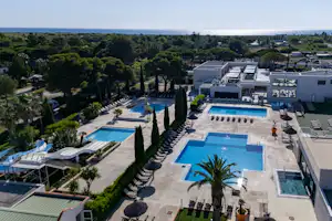 Blick auf mehrere Swimmingpools und Liegen am Campingplatz, umgeben von gepflegten Wegen, Palmen und Bäumen sowie weißen Gebäuden.