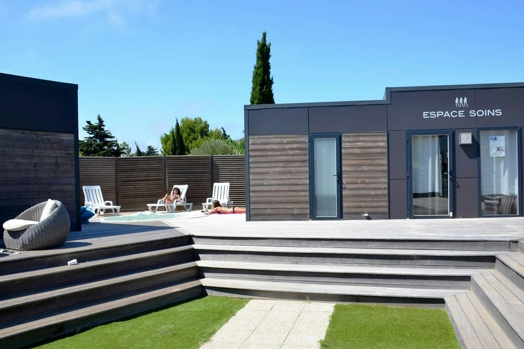 Terrasse mit Poolbereich, mehreren Liegen und Sichtschutz aus Holz vor einem Gebäude mit Eingang „Espace Soins“.