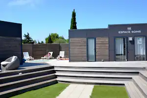 Terrasse mit Poolbereich, mehreren Liegen und Sichtschutz aus Holz vor einem Gebäude mit Eingang „Espace Soins“.