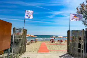 Blick durch ein offenes Tor auf den Strand mit Liegestühlen, Sandweg und zwei Fahnen am Camping-Eingang.