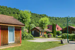 Holzbungalow mit Terrasse, gepflegter Rasenfläche und Kiesweg, umgeben von Bäumen und weiteren Ferienhäusern im Campingbereich.