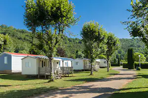 Reihe von Mobilheimen auf einem Campingplatz, mit Holzterrassen, Rasenflächen, Bäumen und einem breiten Weg.