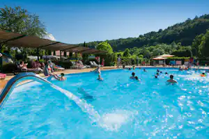 Großes Camping-Schwimmbecken mit Sonnenschirmen und Liegen, Menschen im Wasser, im Hintergrund grüne Hügel und Bäume.