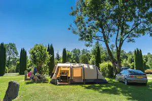 Campingzelt auf einer grünen Wiese, daneben ein Auto und Sitzbereich mit Stühlen unter Bäumen.