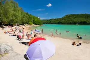 Zelte am Sandstrand am See, viele Badegäste im türkisfarbenen Wasser, Bäume und Hügel im Hintergrund.