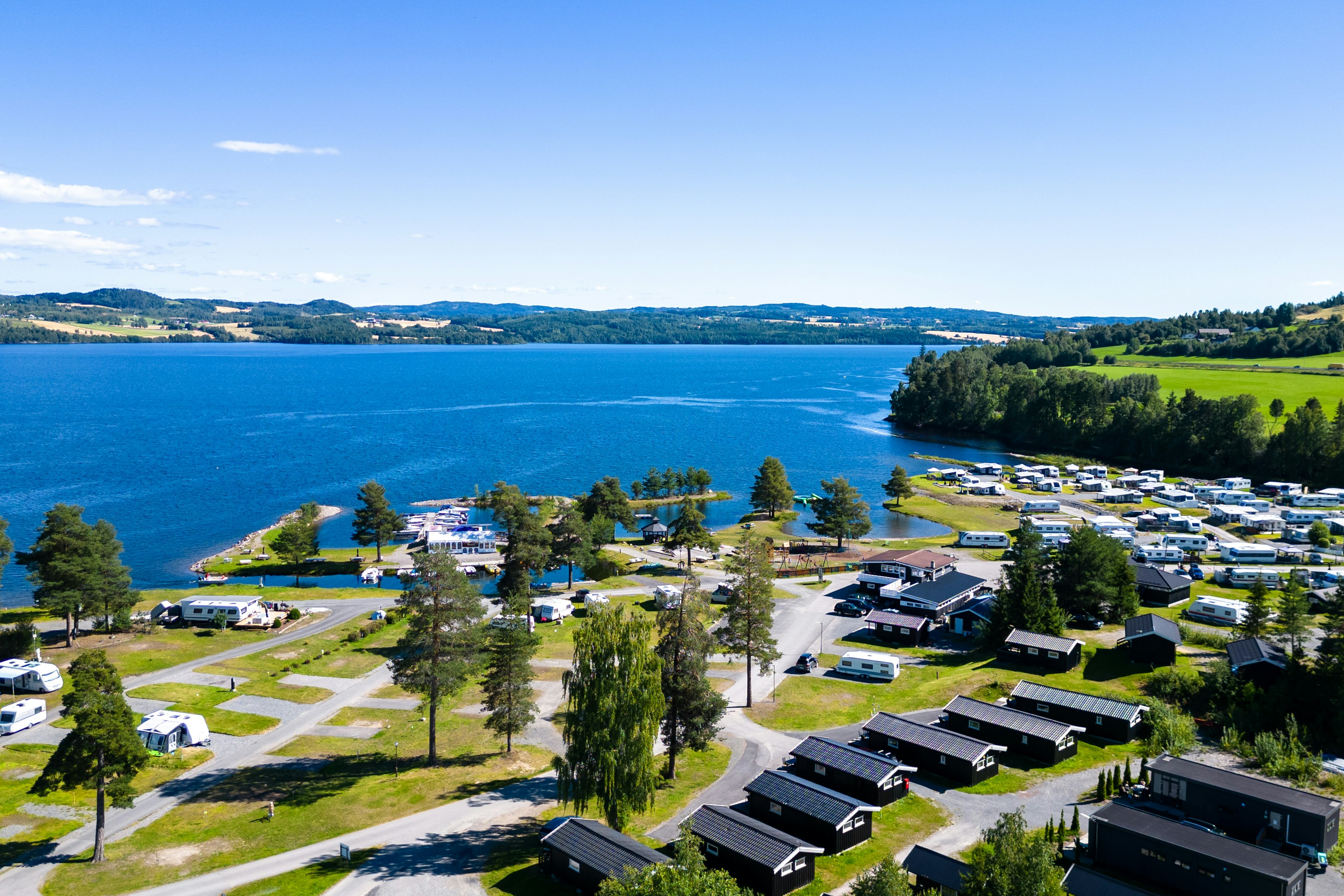Luftaufnahme eines Campingplatzes am See mit Stellplätzen für Wohnmobile, Gebäuden, Wegen und Bäumen am Ufer.