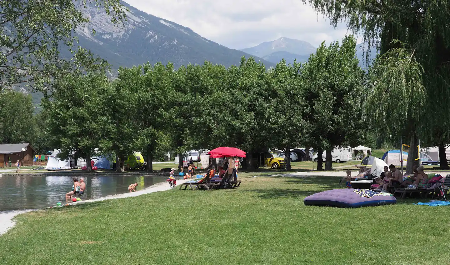 Campingplatz am See mit Rasen, Sitzgruppen und Zelten, im Hintergrund Berge und Bäume.