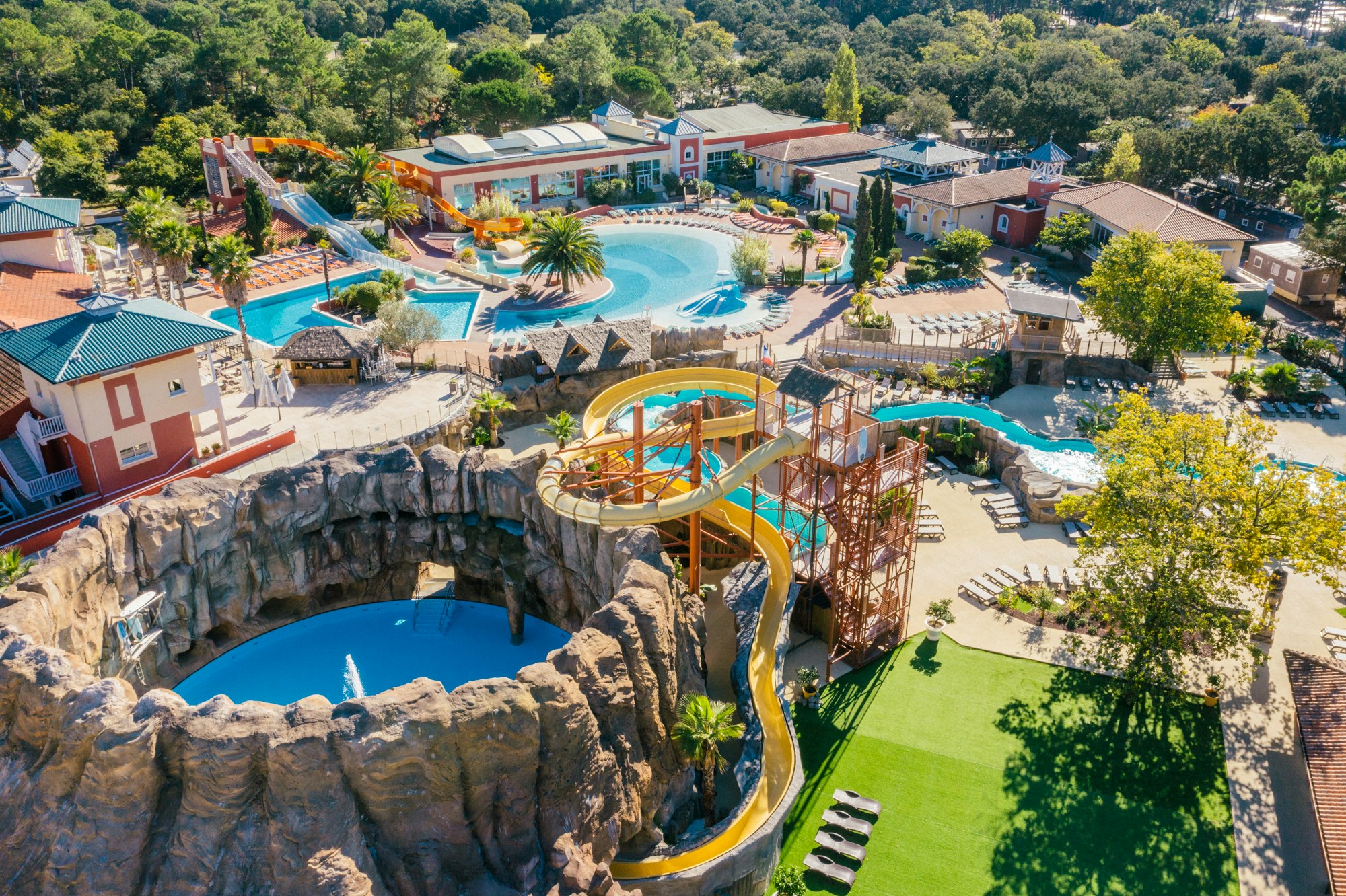 Luftaufnahme eines Campingplatzes mit mehreren Schwimmbecken, Wasser-Rutschen, Liegen und Wegen zwischen Gebäuden und Bäumen.