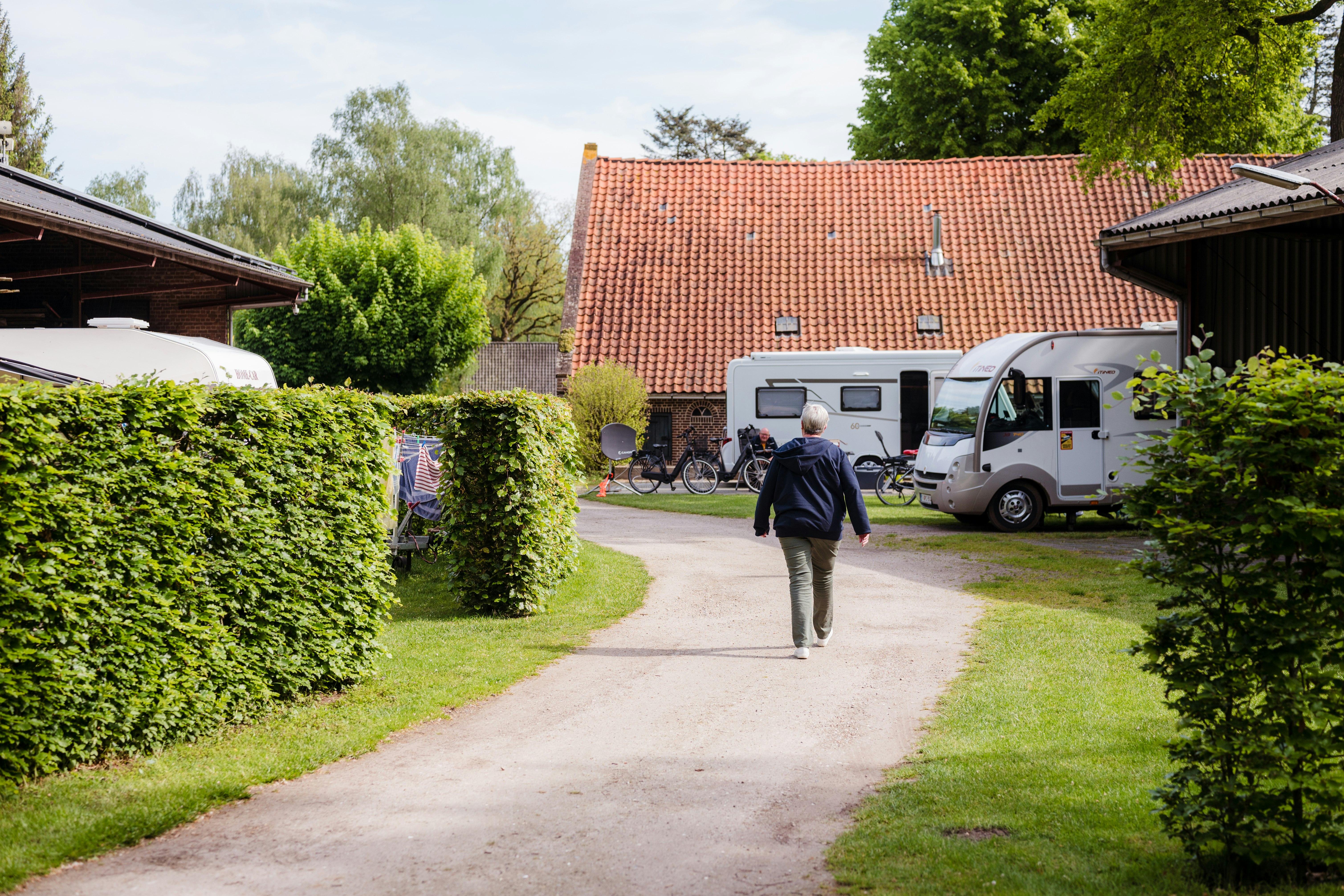Weg im Campingbereich zwischen Hecken, im Hintergrund Wohnmobil und Gebäude mit rotem Ziegeldach.