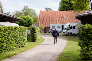 Weg im Campingbereich zwischen Hecken, im Hintergrund Wohnmobil und Gebäude mit rotem Ziegeldach.