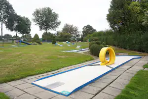 Minigolfbahn auf dem Campingplatz mit gelbem Tor, blauen Bahnen und Rasen, umgeben von Bäumen und Hecken.