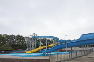 Blaue Wasserrutsche auf einem Campingplatz, mit Geländer und Zaun, im Hintergrund Bäume und ein Spielbereich.