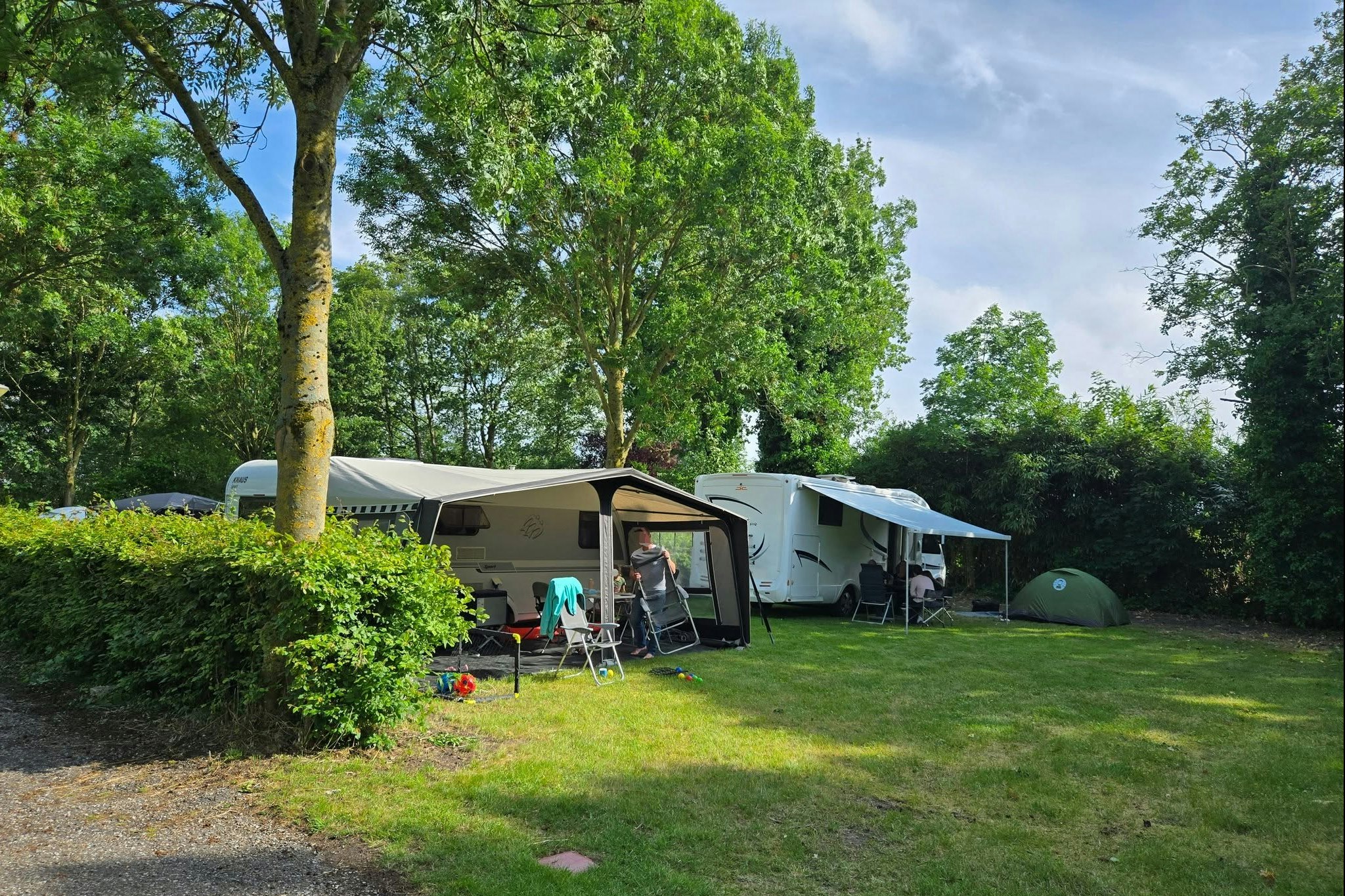 Campingplatz mit Wohnwagen und Vorzelt, Sitzgruppe auf dem Rasen und Hecken im Hintergrund, Bäume und Weg am Bildrand.