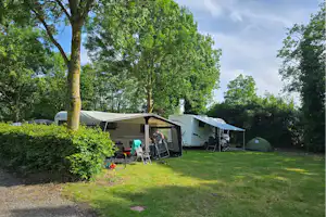 Campingplatz mit Wohnwagen und Vorzelt, Sitzgruppe auf dem Rasen und Hecken im Hintergrund, Bäume und Weg am Bildrand.